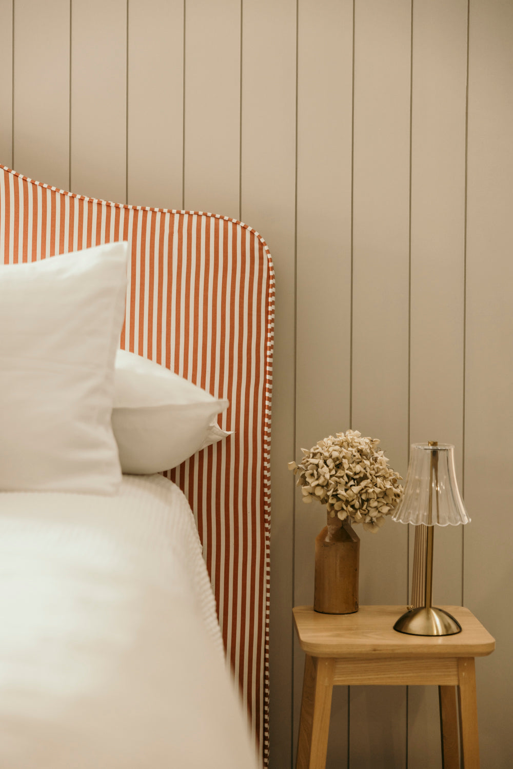 Bed with striped headboard and nightstand with lamp and flowers against a wooden paneled wall.