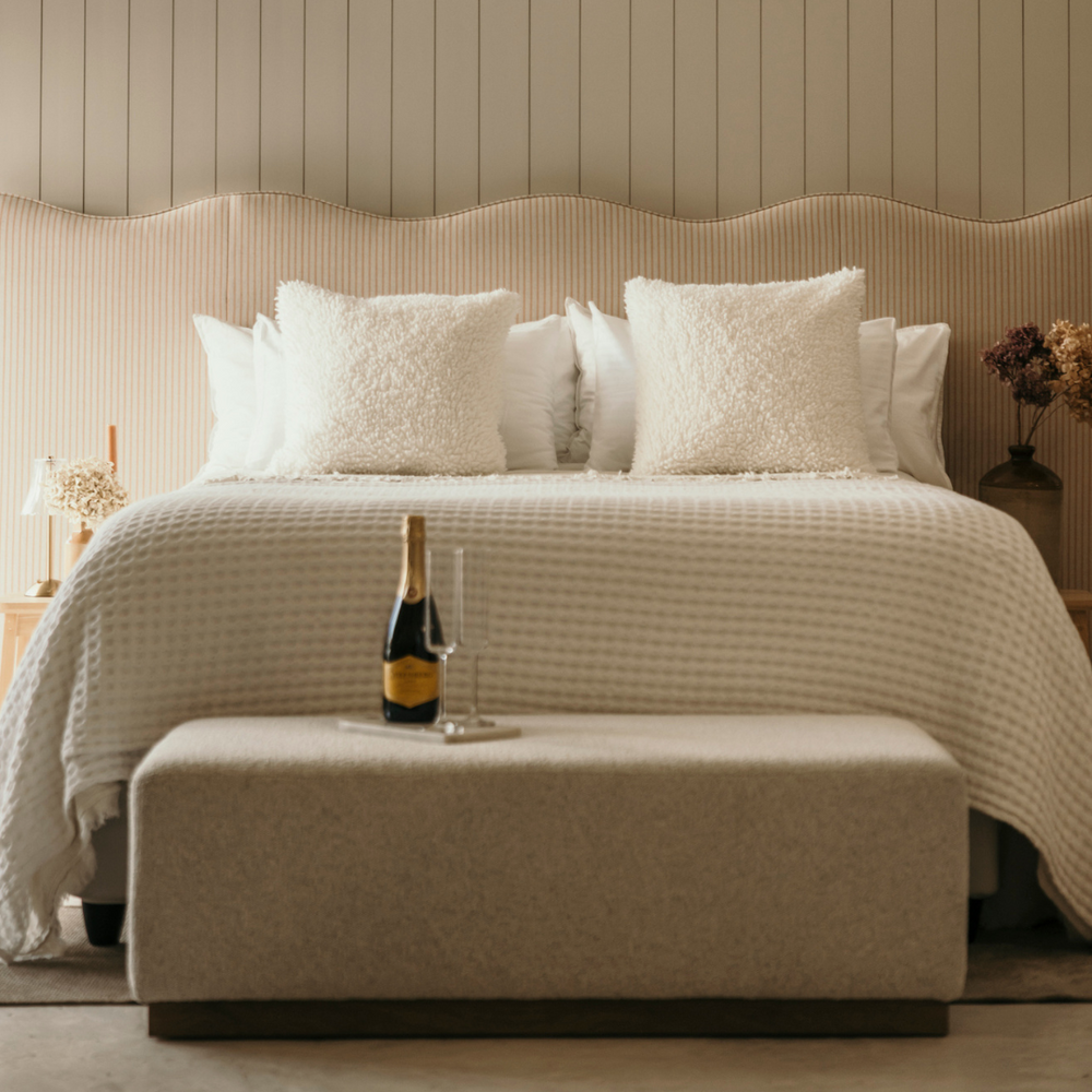 Neatly made bed with decorative pillows and a bottle of champagne on an ottoman in a bedroom setting.