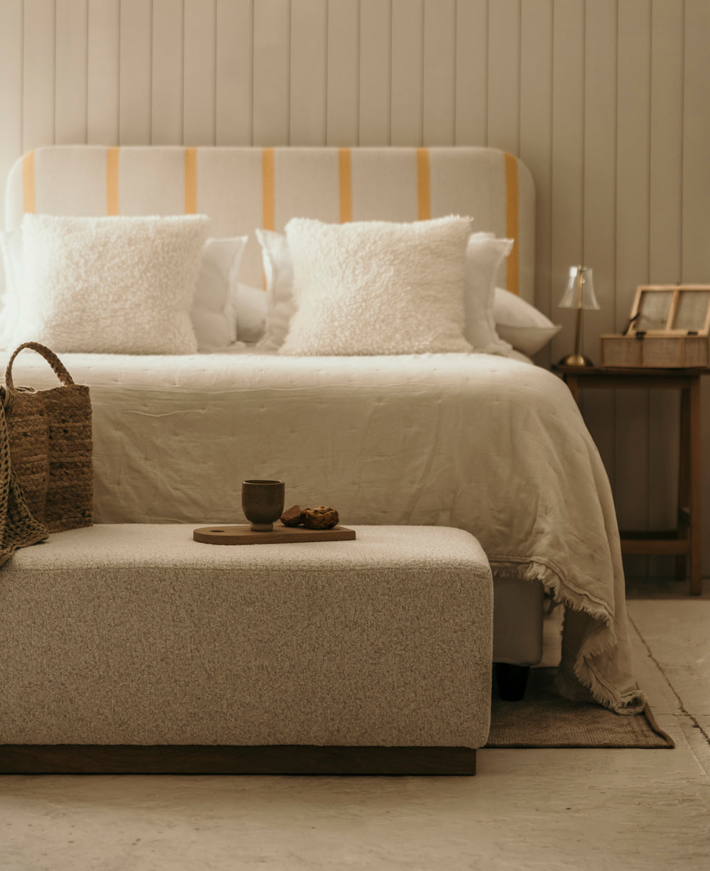 Beige bedroom with a bed and ottoman, featuring soft lighting.
