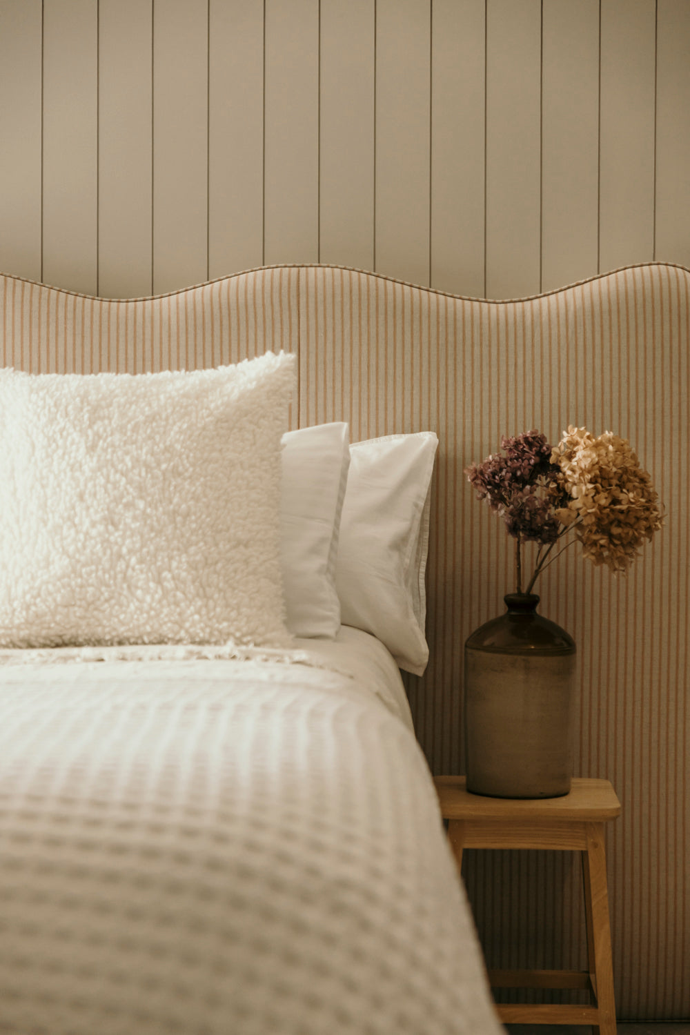 Neatly made bed with decorative pillows and a vase of flowers on a nightstand against a striped wall.
