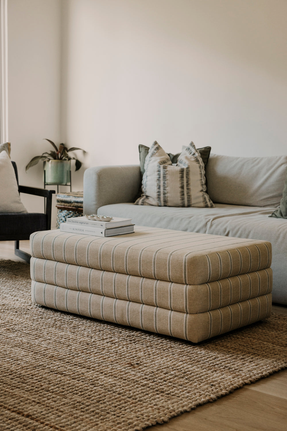 Beige striped ottoman in front of a gray sofa with patterned cushions on a wooden floor.