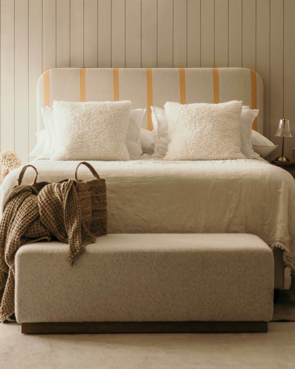 A bedroom with a striped upholstered headboard and a wool bench ottoman at the foot of the bed, from the Holly Wood Design Studio Collection
