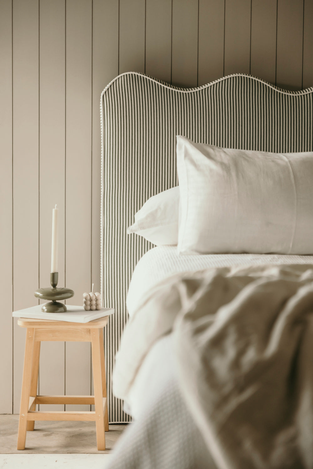 Neatly made bed with white bedding against a striped wall, next to a wooden side table.
