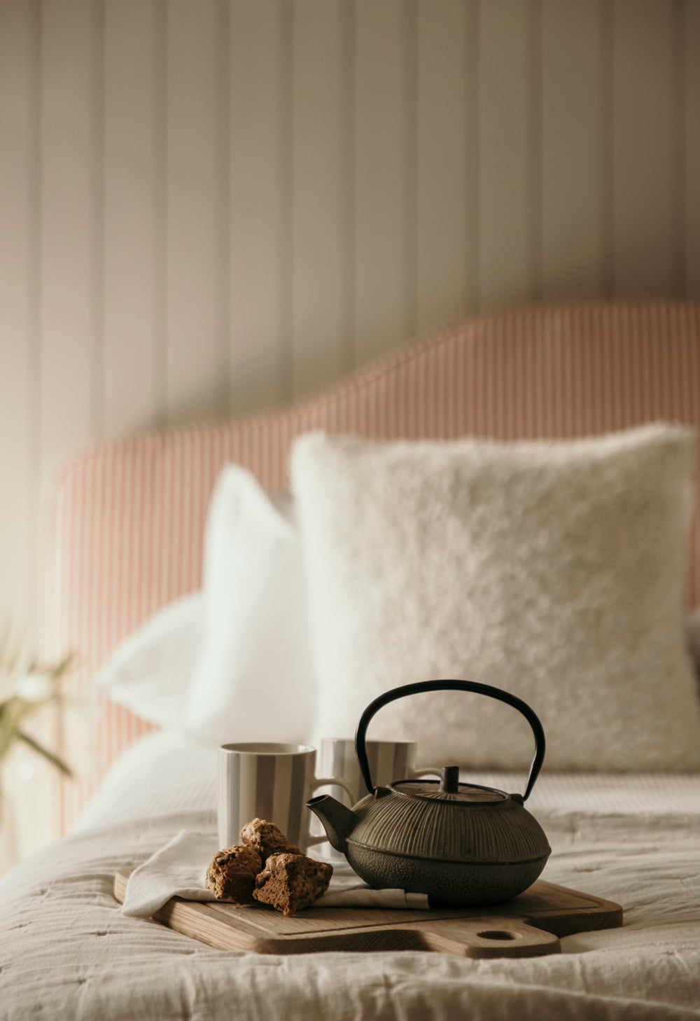 Tea set on a tray with a teapot, cups, and tea leaves on a bed with white pillows and a pink headboard.