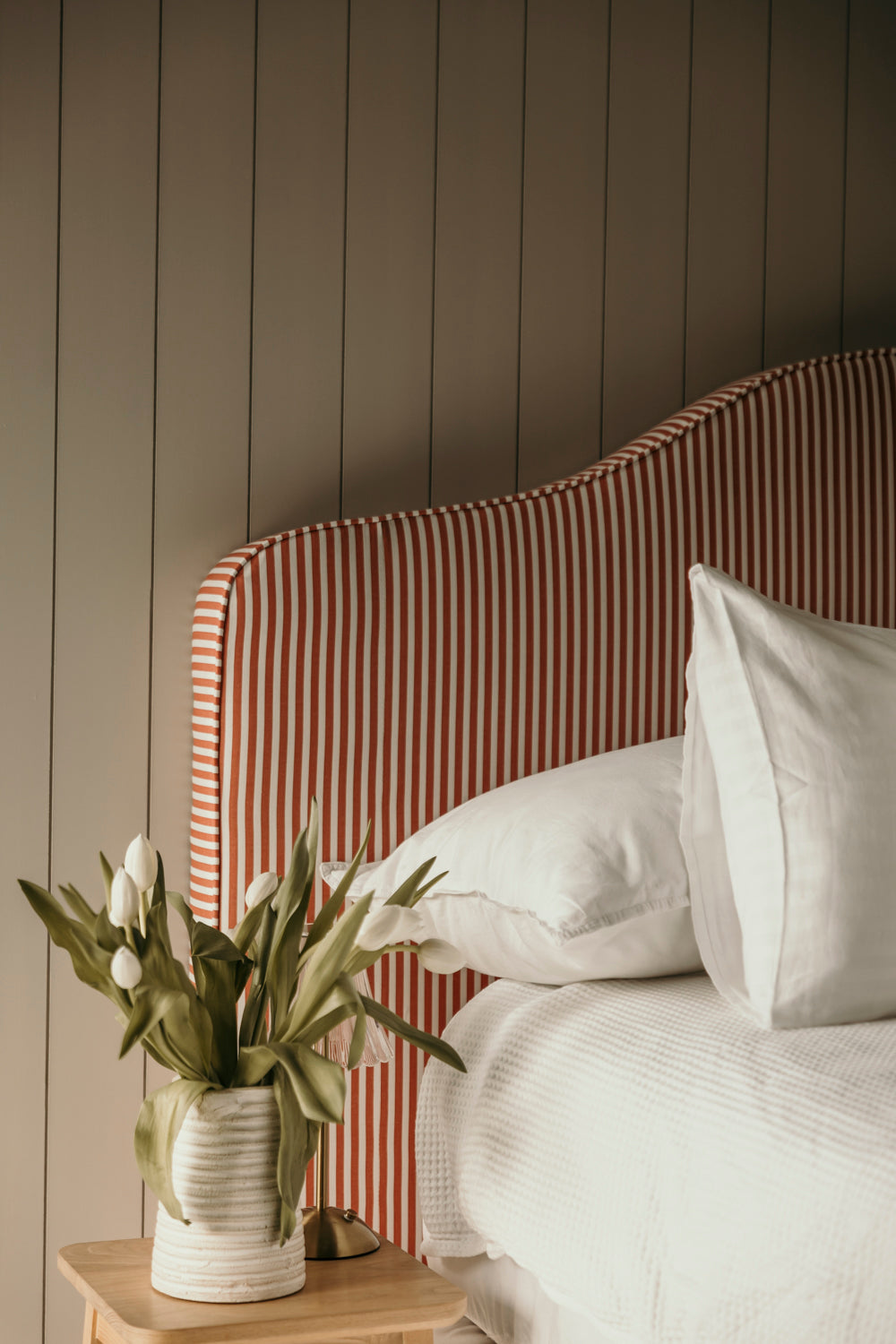 Bed with striped headboard and white pillows, next to a vase with flowers on a wooden side table.