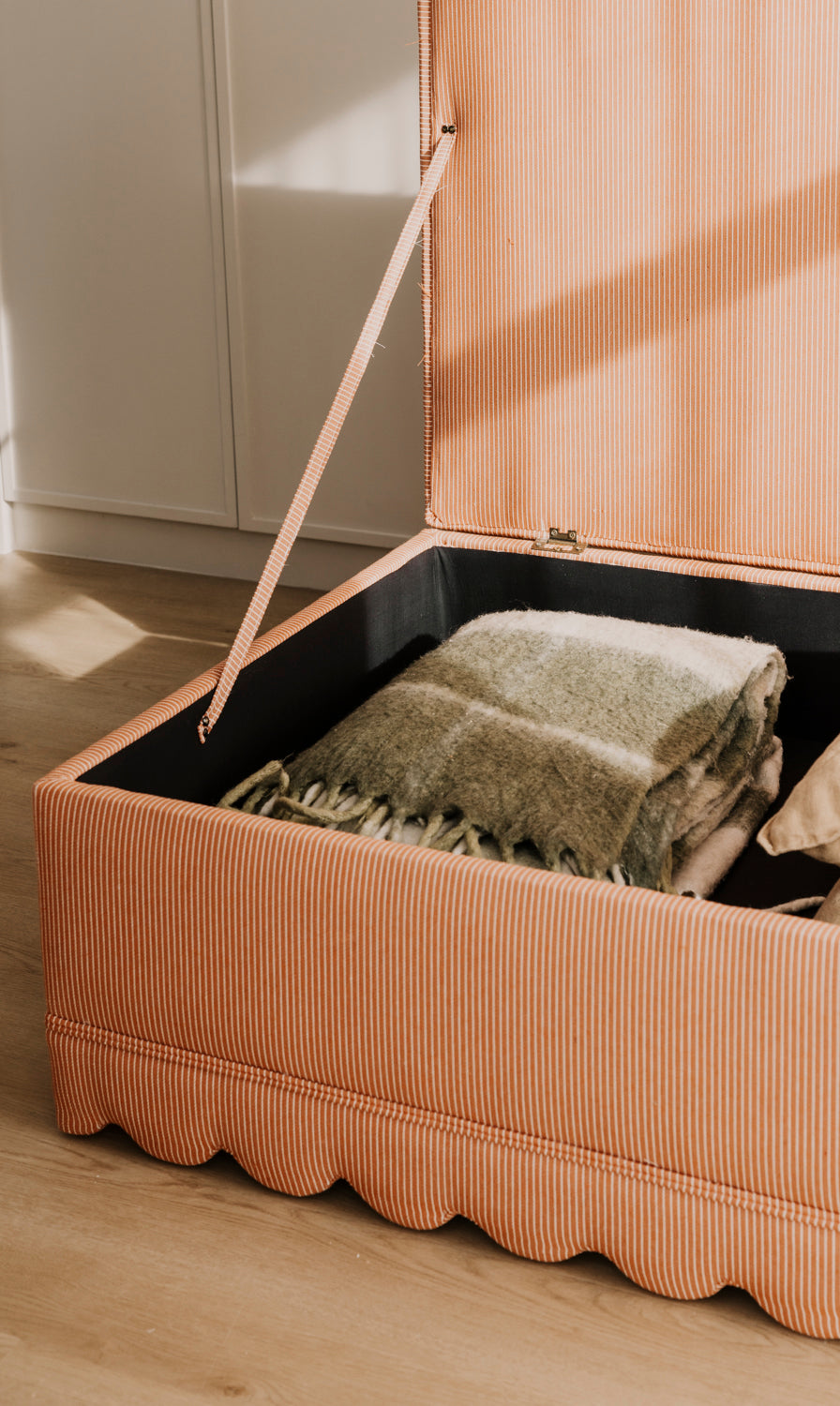 Beige storage ottoman with a blanket inside on a wooden floor.
