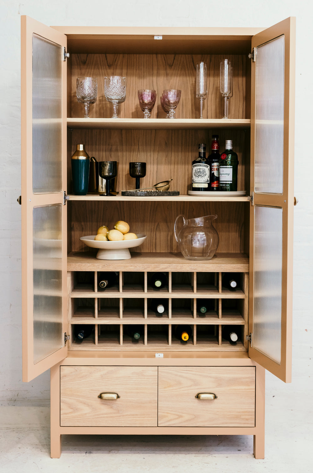 Wooden bar cabinet with open doors displaying bottles, glasses, and fruit on a white background