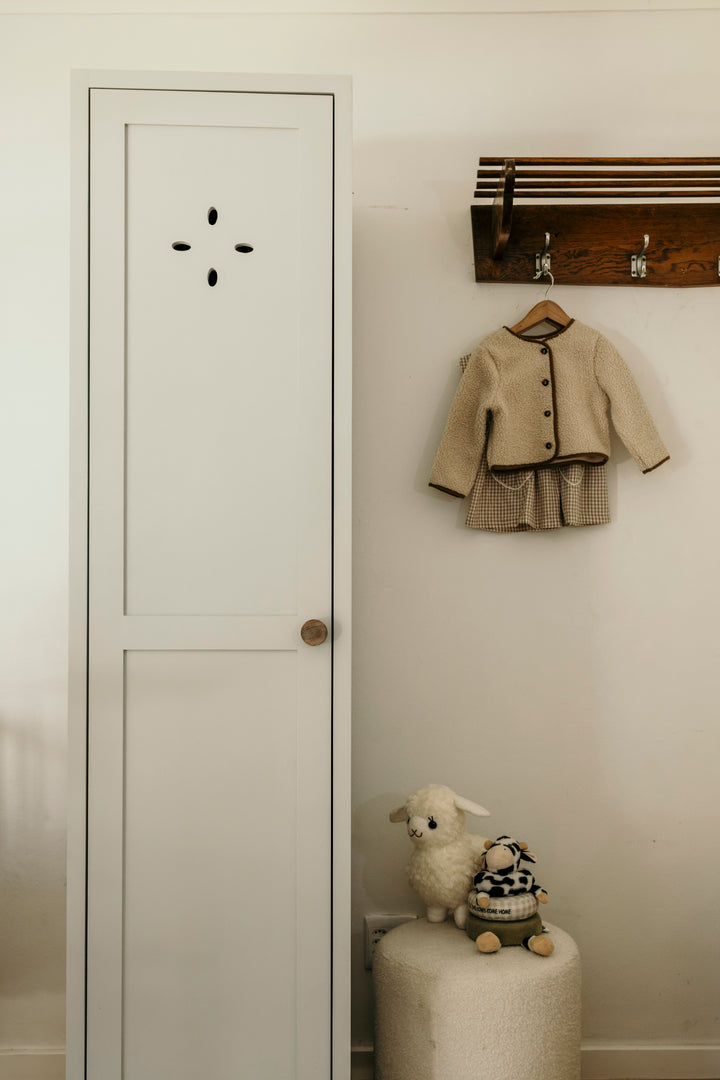 Children's coat hanging on a hook next to a white door with a small sheep plush toy on a pedestal.