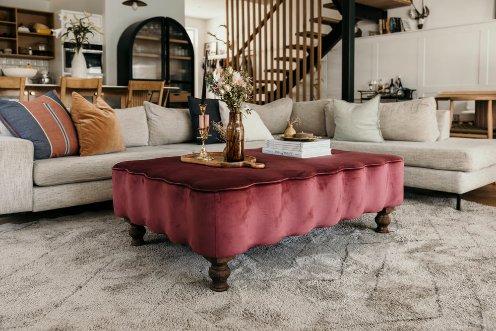 Living room with a burgundy ottoman in the center, surrounded by beige sofas and decorative items.