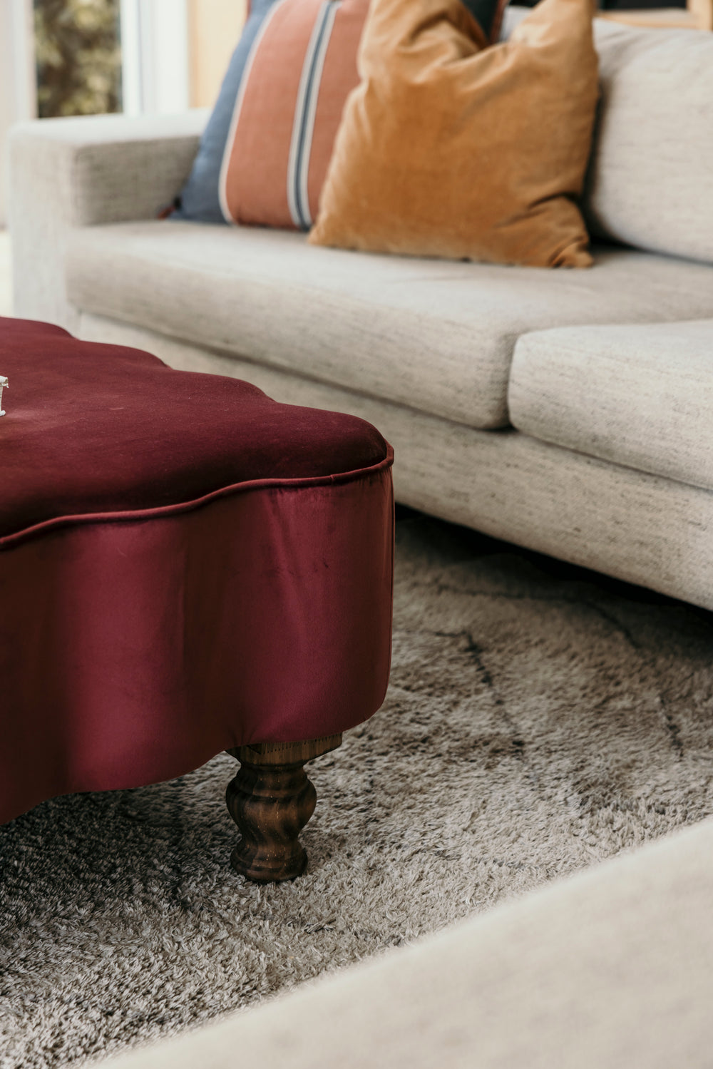 Maroon ottoman in front of a gray sofa with patterned cushions on a textured rug.