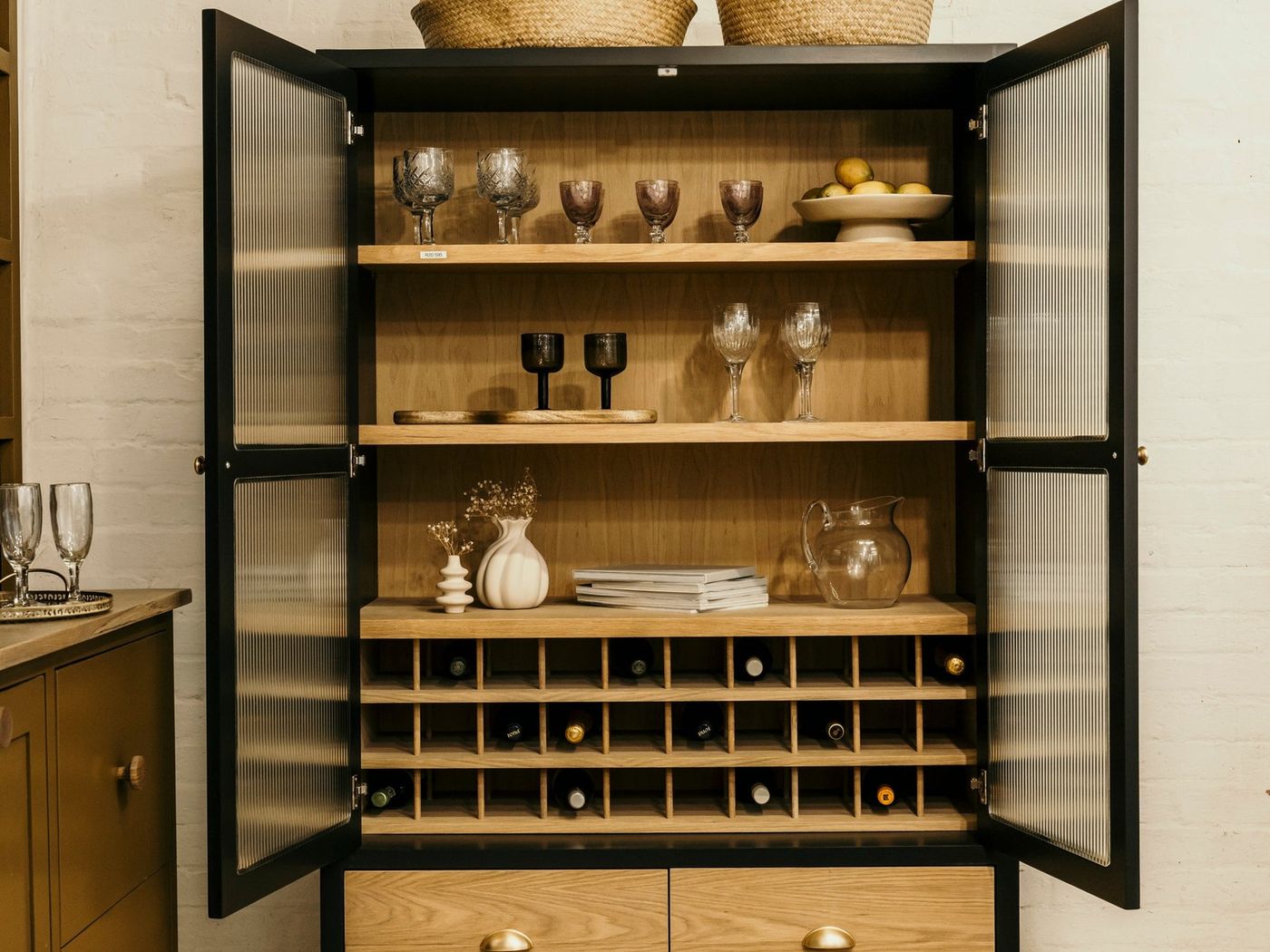 A black and oak glass-fronted Holly Wood cabinet with glassware and a wine rack, in a styled showroom